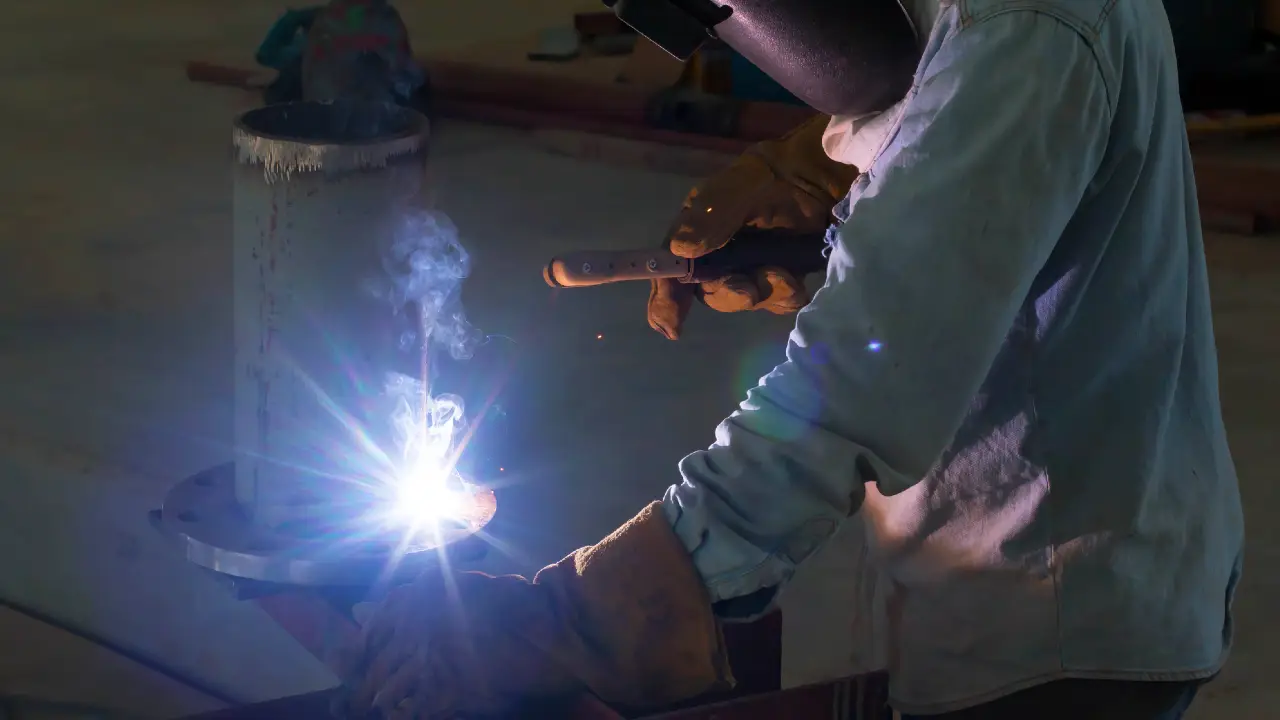 Una persona che indossa guanti protettivi e un casco da saldatore utilizza una torcia da saldatura, producendo scintille luminose e fumo. La scena è illuminata dall'intensa luce bianco-blu del processo di saldatura.
