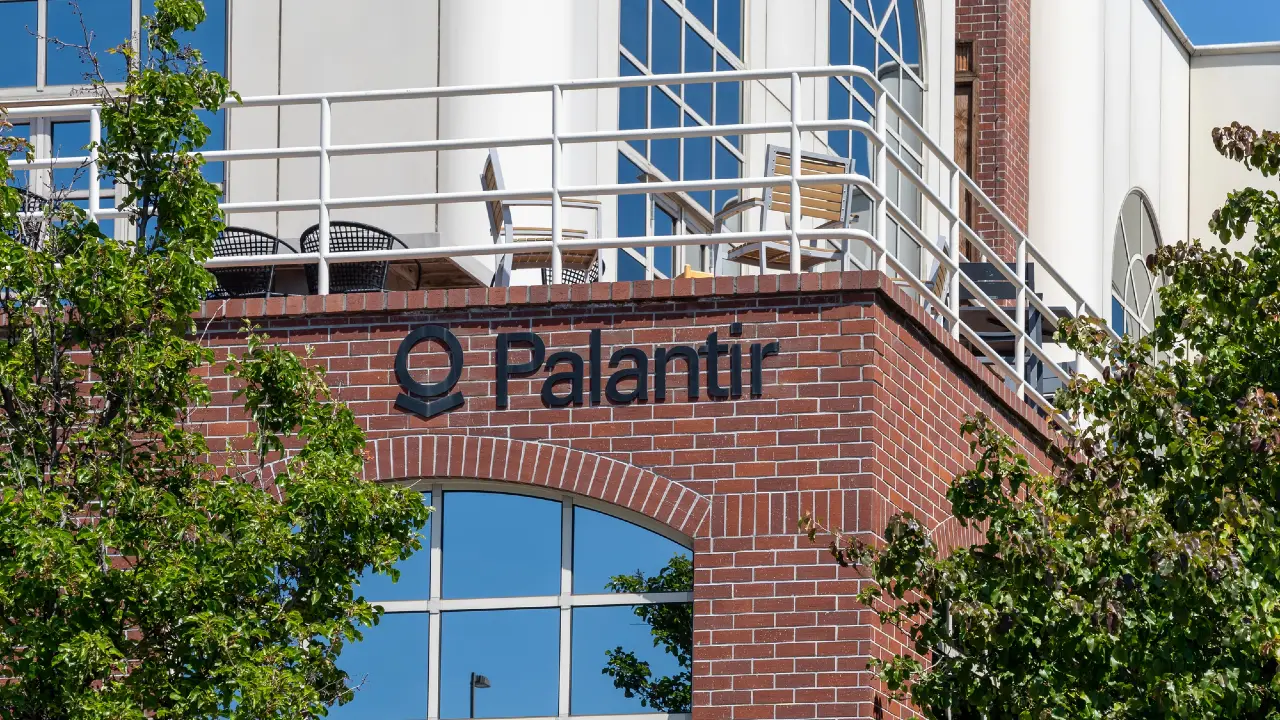 L'esterno di un edificio in mattoni rossi con il logo e il nome di Palantir sul muro, grandi finestre di vetro, ringhiere bianche e alberi che oscurano parzialmente la vista.