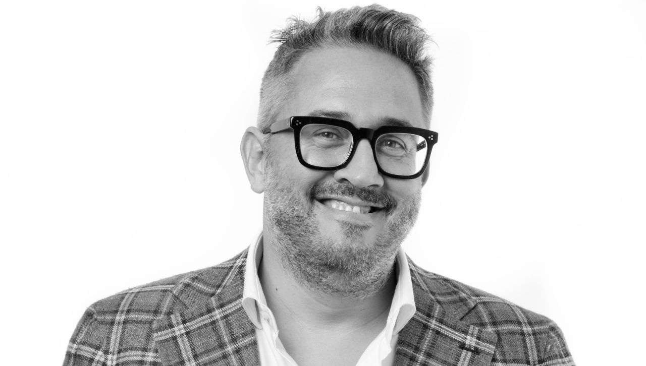 Un uomo sorridente con capelli corti e acconciati, barba e occhiali, che indossa un blazer a quadri su una camicia bianca, davanti a un semplice sfondo bianco.
