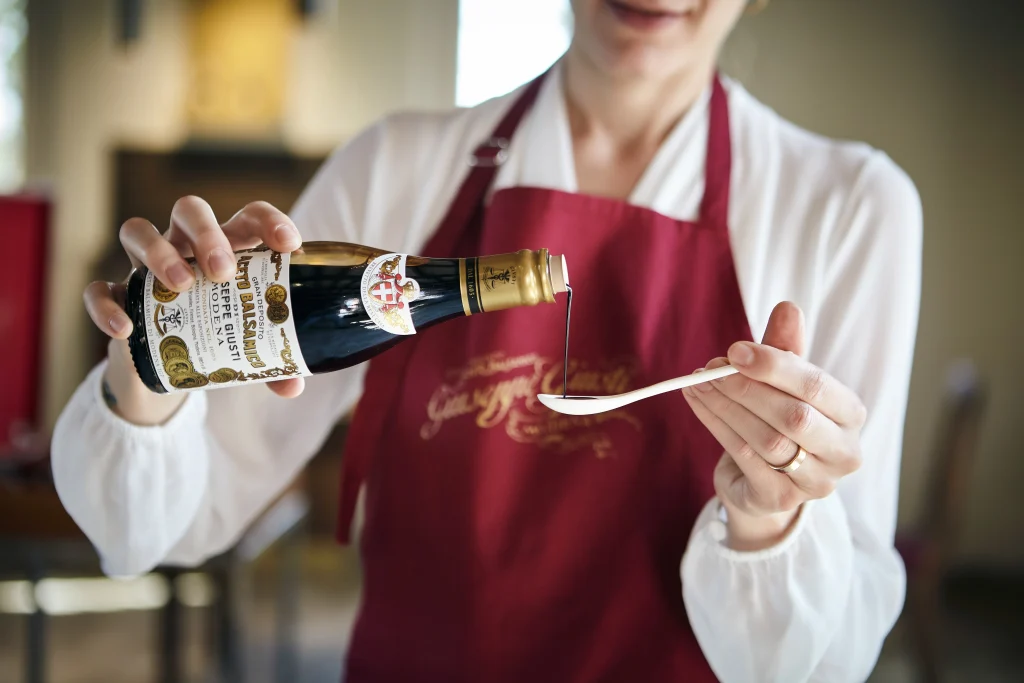 Una persona che indossa un grembiule rosso versa l'aceto balsamico Acetaia Giusti da una bottiglia su un cucchiaio bianco, tenendo entrambi gli oggetti con cura tra le mani.