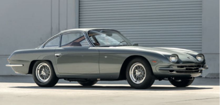 Una classica Lamborghini 400 GT grigia con ruote a raggi metallici è parcheggiata su una superficie di cemento di fronte a un edificio con una grande porta avvolgibile di metallo bianco.