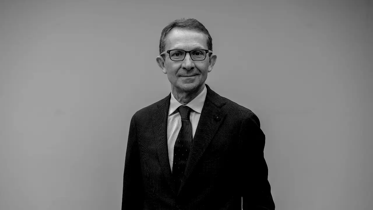 Un uomo in giacca e cravatta, con gli occhiali, si trova su uno sfondo semplice, rivolto verso la macchina fotografica e leggermente sorridente. L'immagine è in bianco e nero.