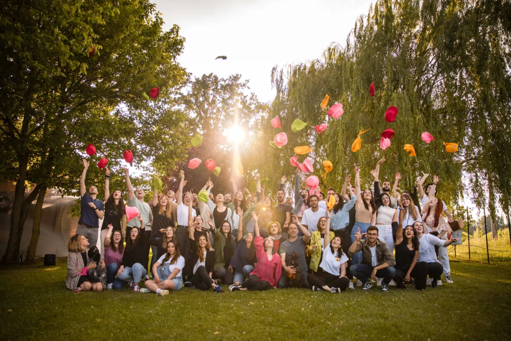 Un folto gruppo di persone si trova sull'erba dell'Acetaia Giusti, applaudendo e lanciando in aria cappelli colorati, con gli alberi e la luce del sole sullo sfondo, creando un'atmosfera celebrativa.
