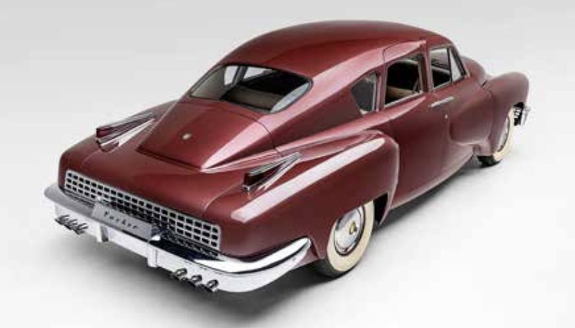 Un Tucker 48 marrone d'epoca visto dalla parte posteriore sinistra, caratterizzato da un design aerodinamico, pinne di coda prominenti, pneumatici bianchi e lunotto posteriore sdoppiato, su uno sfondo chiaro.