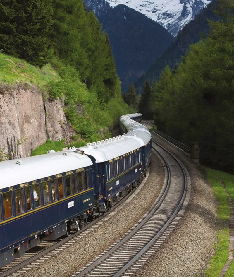 Un treno d'epoca blu e crema, che ricorda l'Orient Express Oggi, viaggia lungo binari curvilinei attraverso una foresta verde e rigogliosa, con montagne e chiazze di neve sullo sfondo.