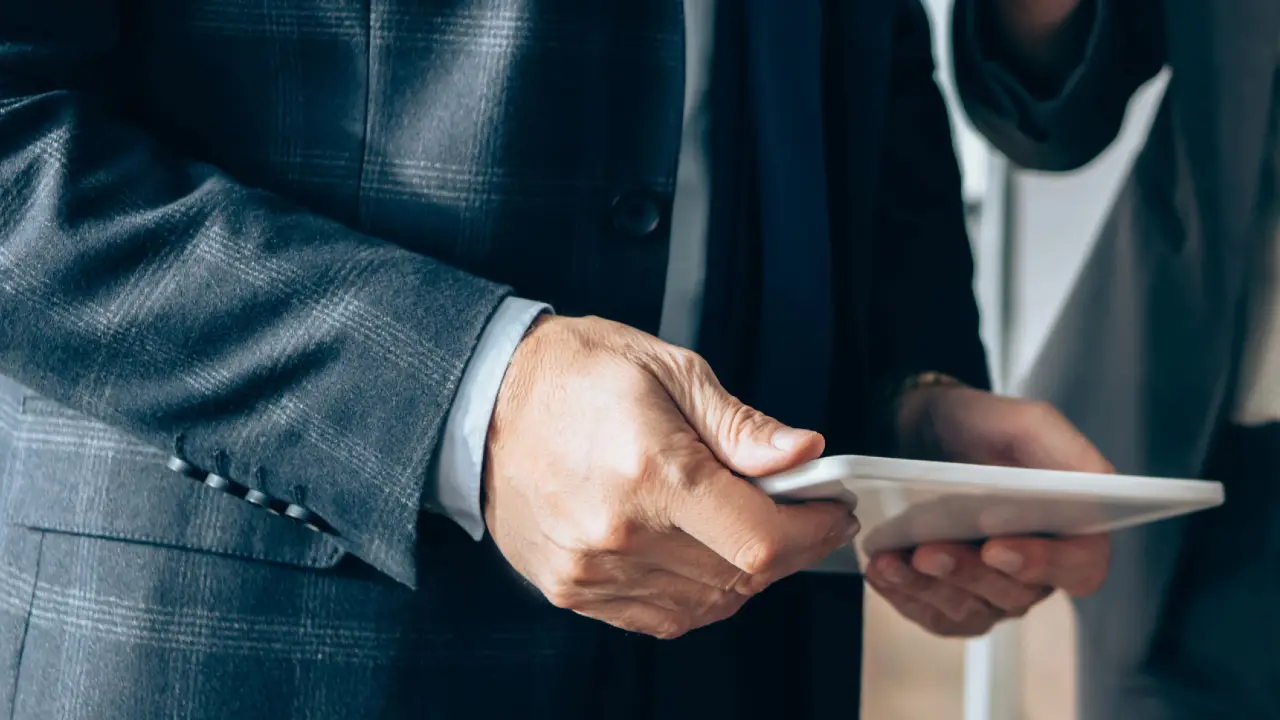 Primo piano di una persona in abito da lavoro che tiene in mano un tablet, con un'altra persona parzialmente visibile accanto. L'attenzione si concentra sulle mani e sul tablet, suggerendo un ambiente professionale o di lavoro.