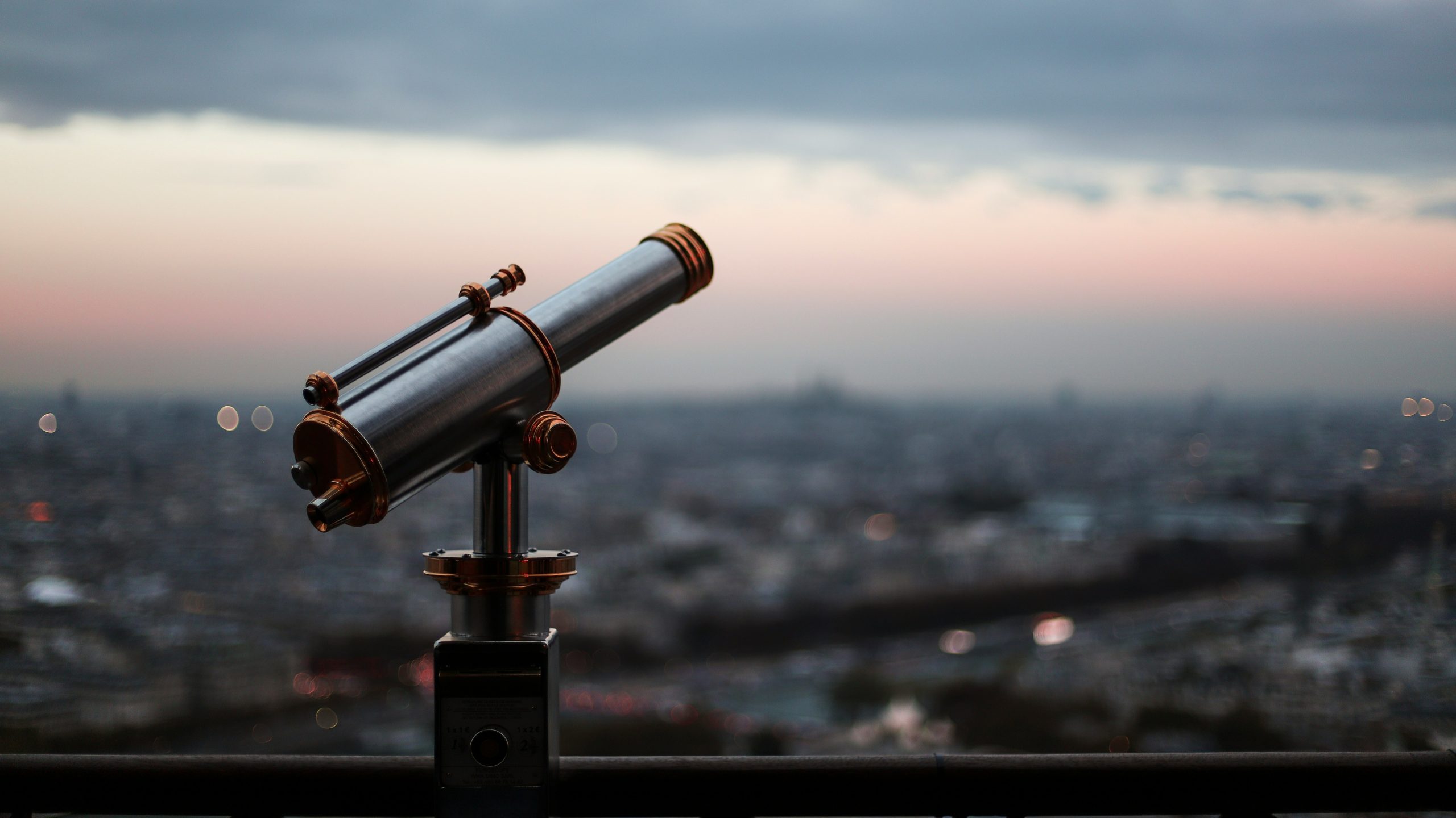Un telescopio pubblico argentato è montato su una piattaforma e si affaccia su un paesaggio urbano al crepuscolo con un cielo rosa e blu sullo sfondo. La città sottostante è sfocata.