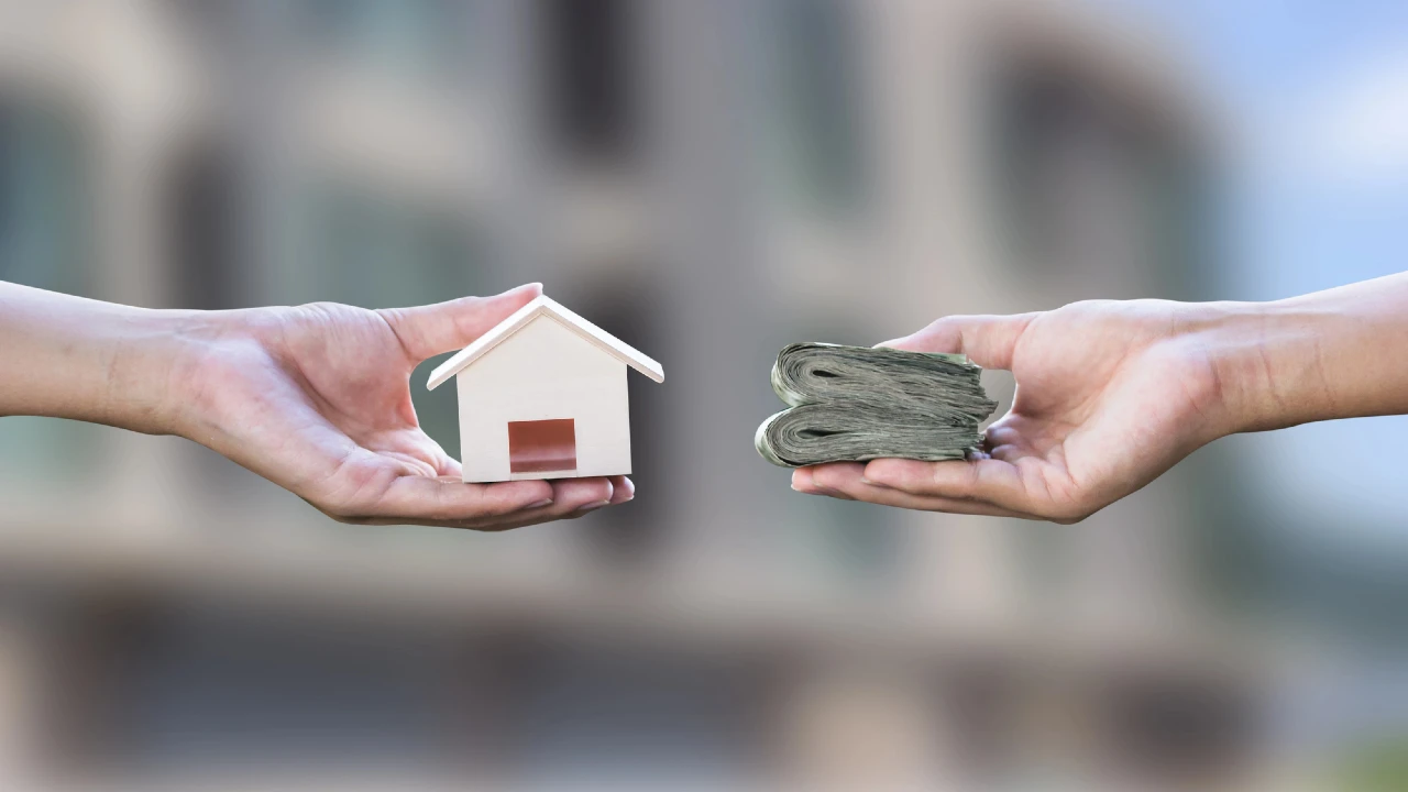 La mano di una persona che regge un modellino di casa è tesa verso un'altra mano con una pila di contanti, a simboleggiare un mutuo o una transazione immobiliare. Lo sfondo sfocato mantiene l'attenzione sullo scambio.