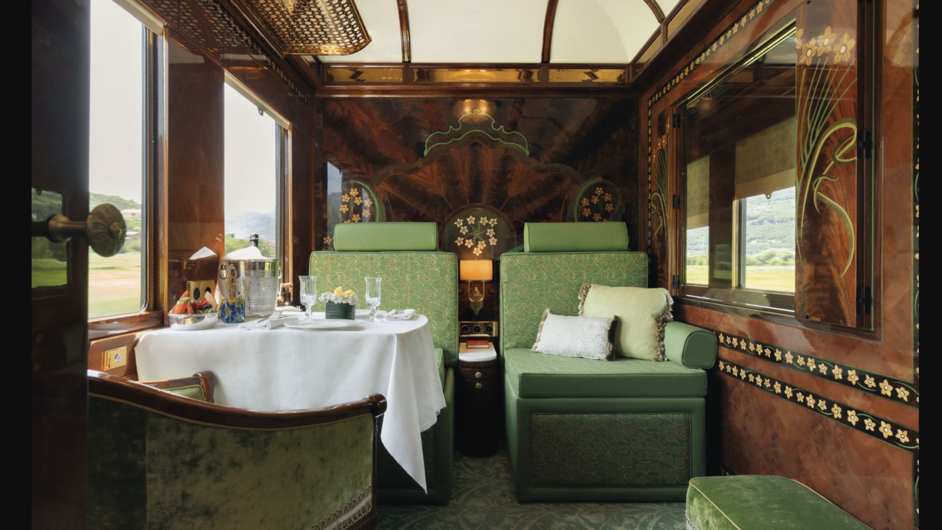 Lussuosa cabina di un treno d'epoca che ricorda l'Orient Express Oggi, con pannelli in legno decorati, sedili imbottiti di verde e un tavolo per due persone con biancheria bianca, fiori e bicchieri. Un'ampia finestra incornicia una vista panoramica sulla campagna.