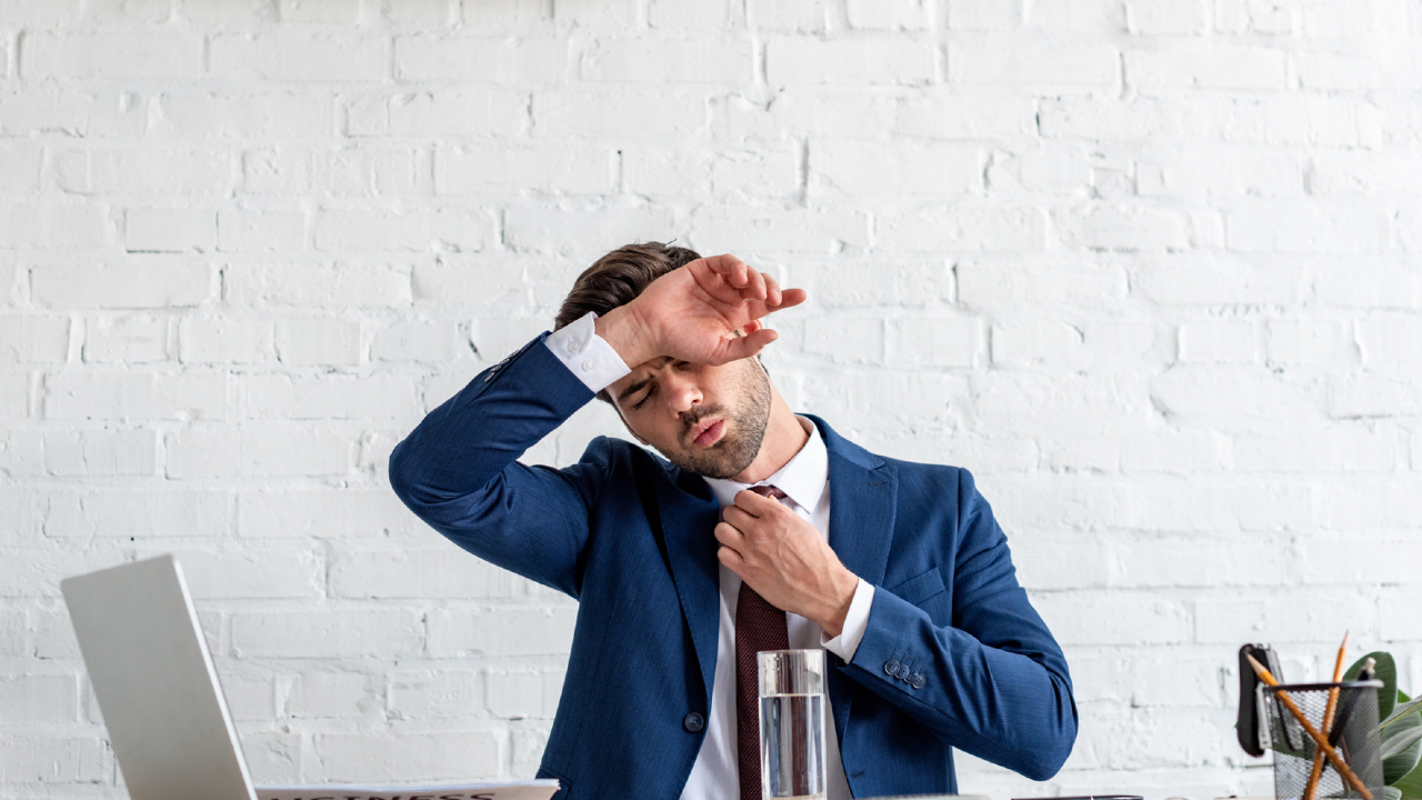 Un uomo in abito blu siede alla scrivania, allentando la cravatta e asciugandosi la fronte, con un'aria stanca o stressata. Sulla scrivania di fronte a lui ci sono un bicchiere d'acqua, un computer portatile e dei fogli.