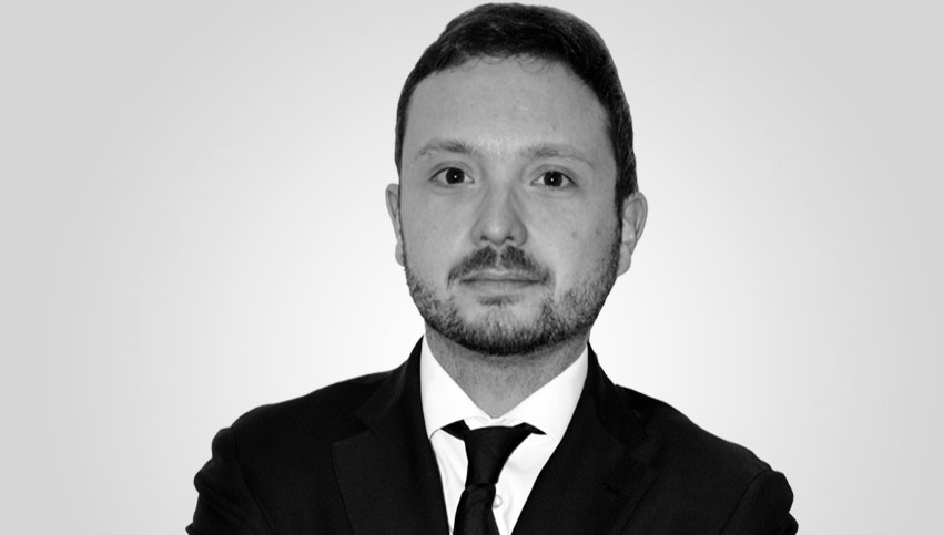 Un uomo con capelli corti e scuri e barba curata indossa giacca e cravatta. È rivolto verso la telecamera con un'espressione neutra. Lo sfondo è semplice e di colore chiaro.