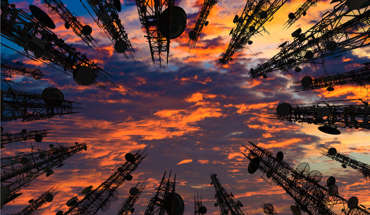 Torri cellulari con numerose antenne e parabole si stagliano su un cielo drammatico al tramonto, pieno di nuvole arancioni, gialle e blu. Le torri sono posizionate intorno ai bordi e puntano verso l'interno.