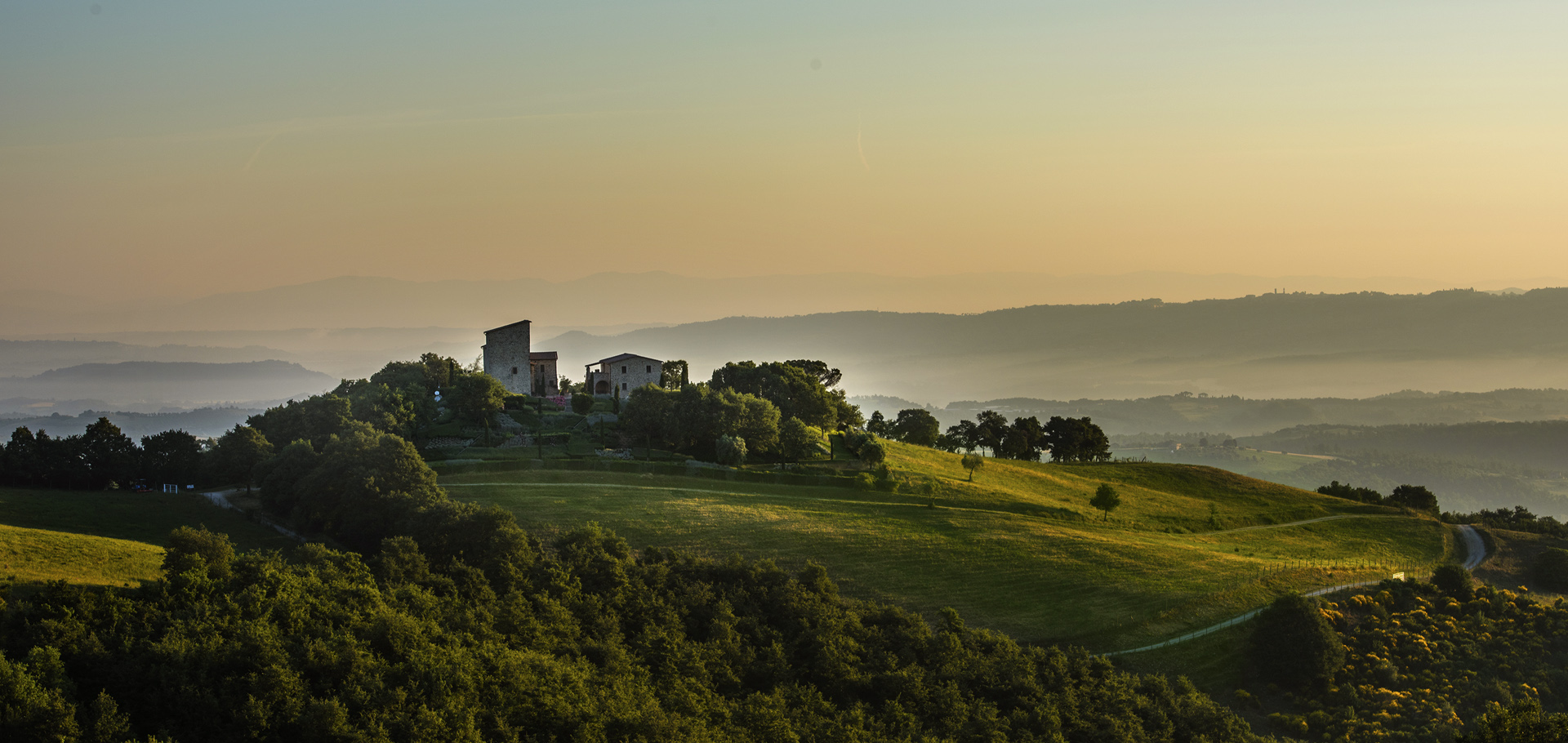 A hilltop villa surrounded by trees and lush green fields, with rolling hills and mountains fading into the misty, golden horizon at sunrise or sunset.