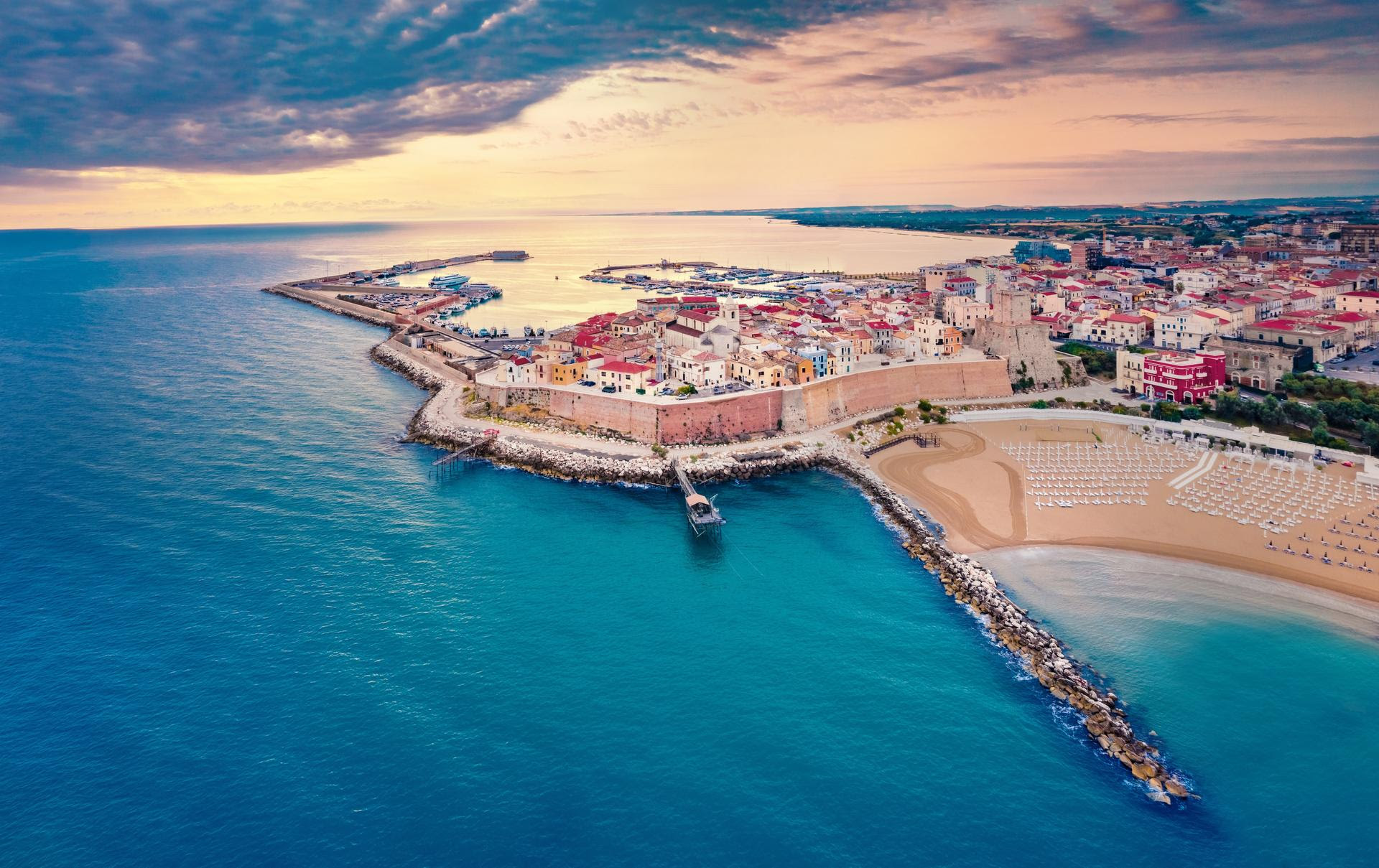 Veduta aerea di una città costiera con edifici colorati, una spiaggia sabbiosa con ombrelloni, frangiflutti rocciosi e un mare azzurro e calmo sotto un drammatico cielo parzialmente nuvoloso al tramonto.