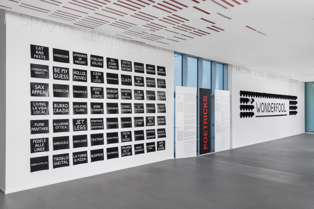 Una galleria d'arte moderna dalle pareti bianche espone griglie di opere tipografiche in bianco e nero con varie frasi. Sostenuta da Leonardo Assicurazioni arte, la parola "WONDERFOOL" campeggia a caratteri neri e decisi sulla parete di fondo.