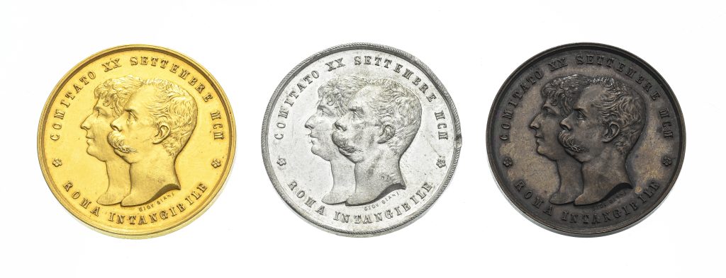 Tre medaglie commemorative di Savoia - d'oro, d'argento e di bronzo - raffigurano due profili maschili rivolti verso sinistra, con iscrizioni in italiano intorno ai bordi. Le monete sono uniformemente distanziate su uno sfondo bianco.