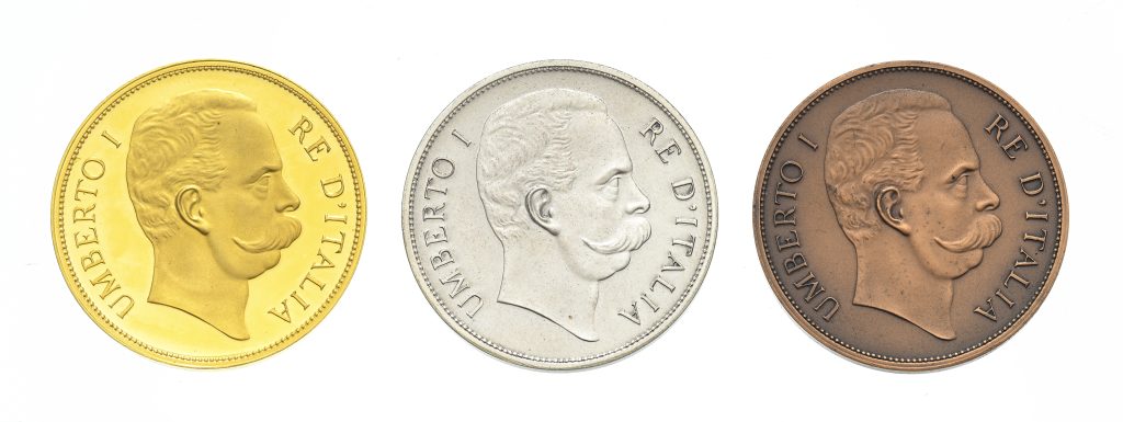 Tre monete italiane con un ritratto di profilo di Umberto I rivolto verso destra, con la scritta "UMBERTO I RE D'ITALIA" lungo il bordo; queste medaglie Savoia sono di colore oro, argento e bronzo.