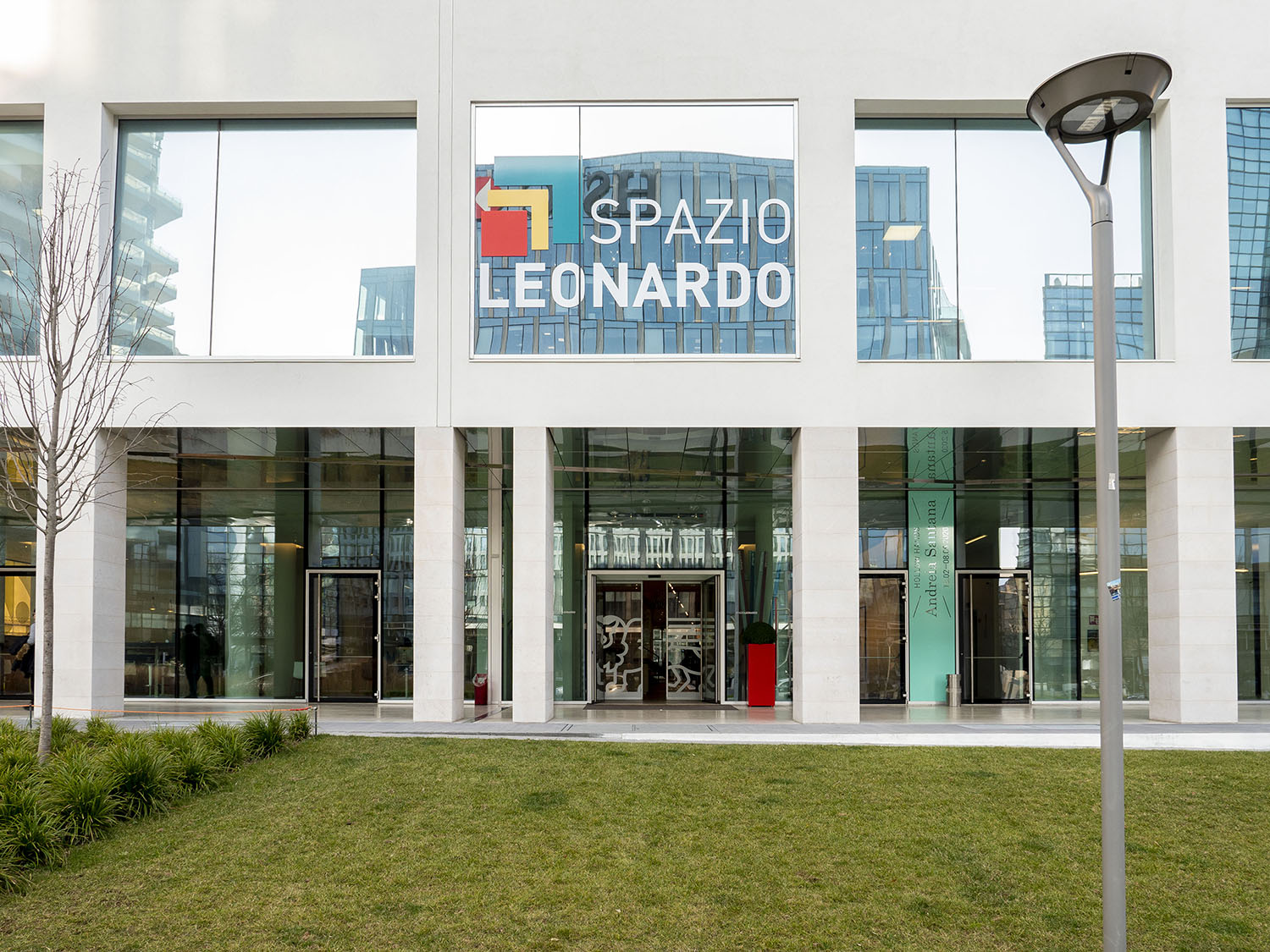 L'ingresso di un edificio moderno con grandi vetrate e un'insegna sopra la quale si legge "Spazio Leonardo". C'è un prato verde, un lampione e un paesaggio che riflette lo spirito creativo di Leonardo Assicurazioni arte.