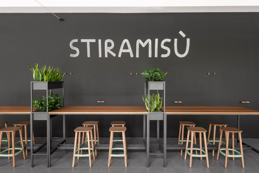 Una caffetteria moderna con tavoli e sgabelli alti in legno, piante verdi in vaso su scaffali e una parete scura con la scritta "STIRAMISÙ" a grandi lettere bianche che unisce il comfort allo stile artistico di Leonardo Assicurazioni.