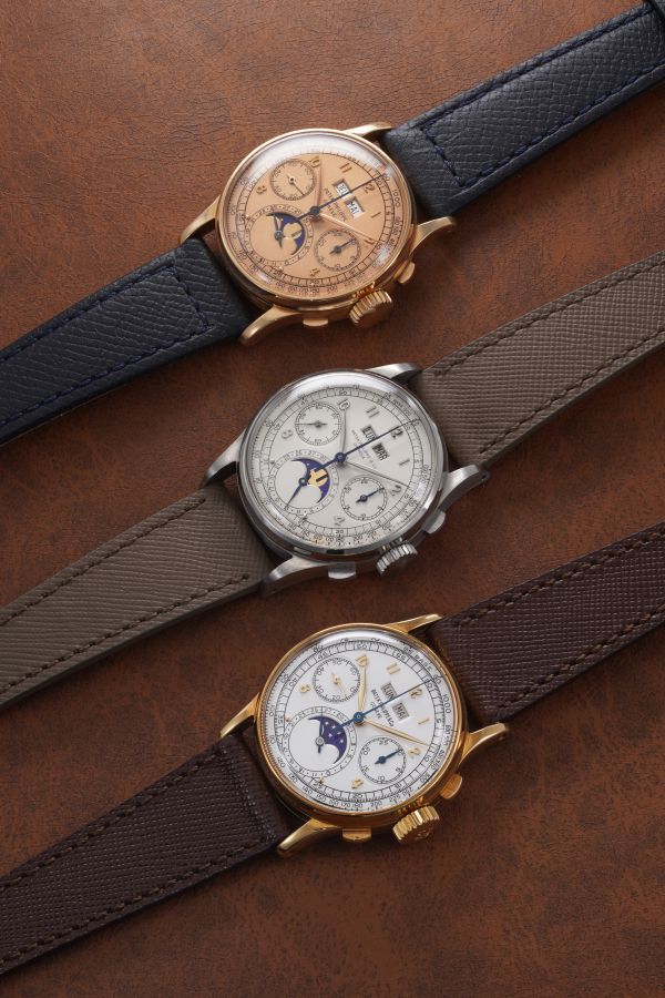 Tre orologi da polso di lusso, ispirati all'iconico Patek Philippe 1518 asta, con cinturini in pelle appoggiati verticalmente su una superficie in pelle marrone. Ciascuno di essi presenta quadranti multipli e un indicatore delle fasi lunari in casse d'oro, d'argento e d'oro.