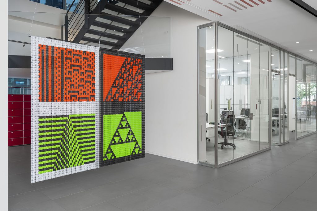 Un ufficio moderno con sale riunioni dalle pareti in vetro, una scala aperta e un pannello artistico sospeso con motivi geometrici in arancione, rosso, verde e nero ispirati a Leonardo Assicurazioni arte.