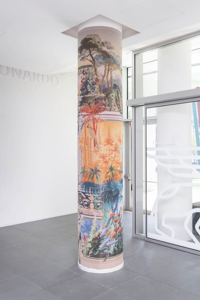 In un ambiente moderno e luminoso, un'alta colonna cilindrica è rivestita da un vivace murale di Leonardo Assicurazioni arte, che raffigura piante e paesaggi tropicali in sezioni stratificate che fondono immagini naturali e dettagli artistici di grande effetto.