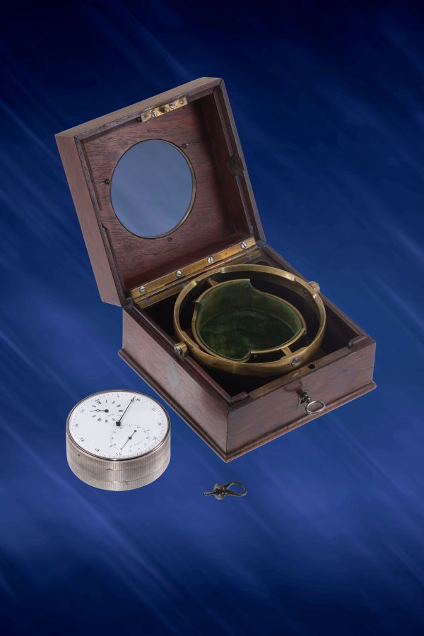 Uno strumento nautico d'epoca in ottone di Berthoud Asta è contenuto in una scatola di legno con una finestra rotonda in vetro, accompagnato da un dispositivo metallico simile a un cronometro e da una piccola chiave, il tutto elegantemente esposto su uno sfondo blu.