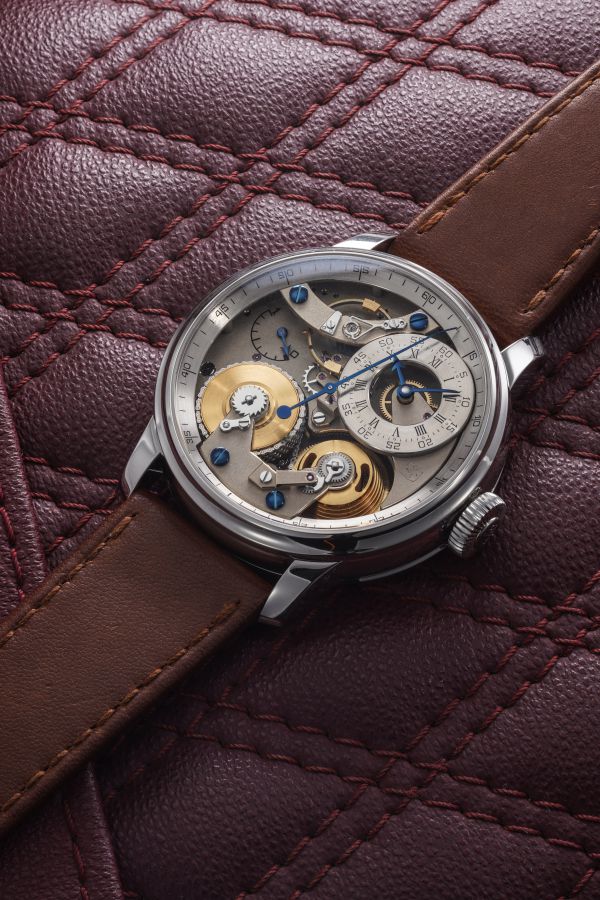 Un orologio da polso meccanico con ingranaggi a vista e cinturino in pelle marrone poggia su una superficie trapuntata in pelle bordeaux, il cui design ricorda la classica lavorazione Berthoud Asta.