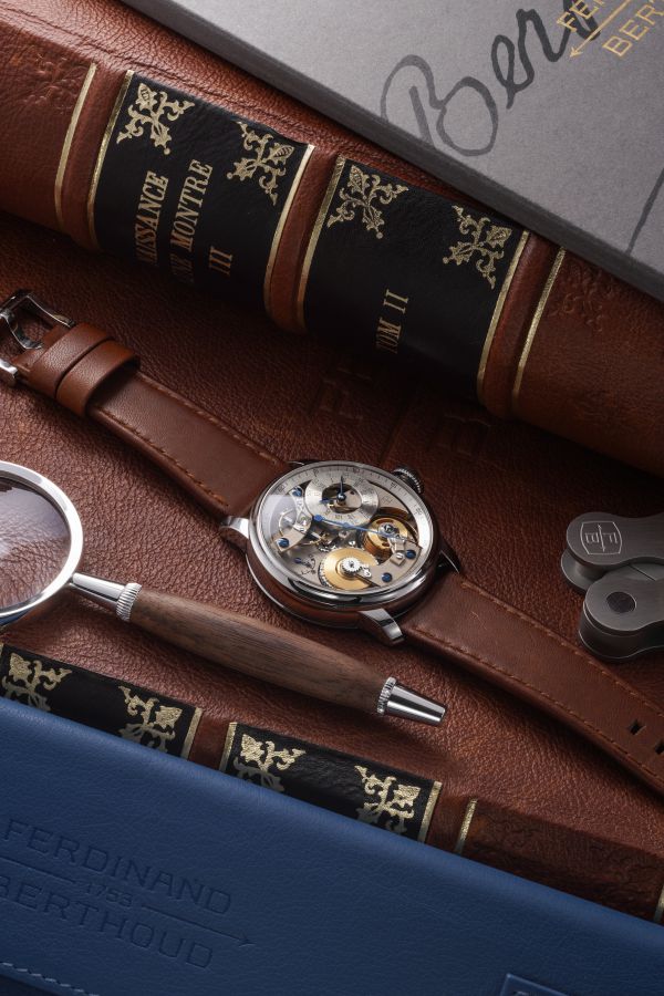 Un orologio da polso meccanico con un intricato movimento visibile giace su un cinturino di pelle marrone, circondato da libri d'epoca, una lente d'ingrandimento, una penna, chiavi di lusso e una cartella blu con l'etichetta "Ferdinand Berthoud Asta".