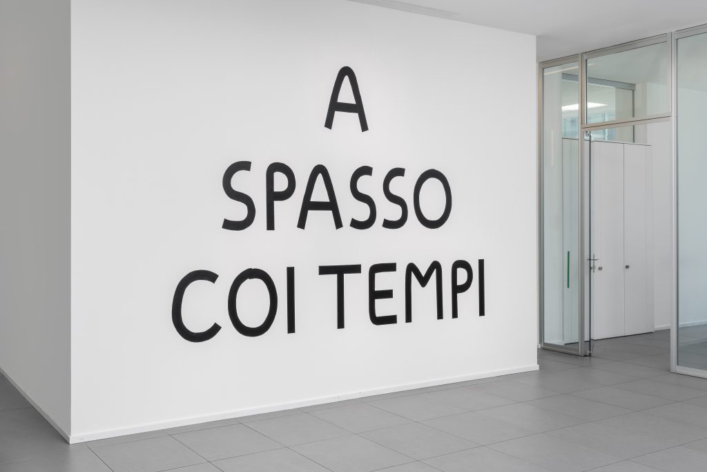 Un testo nero su una parete bianca recita "A SPASSO COI TEMPI" in caratteri grandi e distanziati, riflettendo l'estetica moderna e minimalista di Leonardo Assicurazioni arte con pareti divisorie in vetro e pavimenti grigi.
