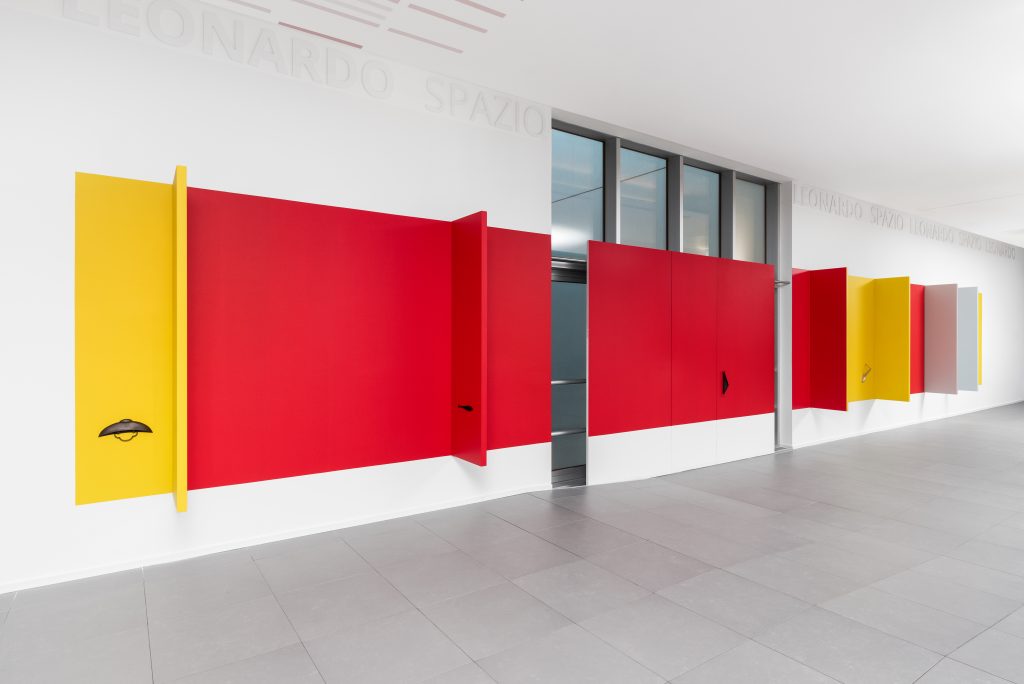 Grande opera d'arte geometrica a parete con pannelli rossi e gialli alternati a elementi scultorei, installata lungo un corridoio bianco con pavimento grigio e porte in vetro. Il sottile testo "LEONARDO SPAZIO" fa riferimento alla Leonardo Assicurazioni arte di cui sopra.
