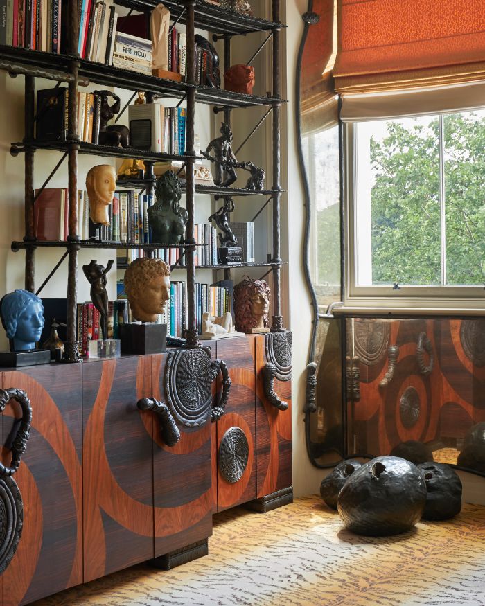 Una stanza elegante ispirata a Pauline Karpidas presenta una libreria piena di libri, busti e sculture sopra un mobile in legno a motivi. Grandi finestre con tende arancioni lasciano entrare la luce naturale e due sgabelli di forma organica poggiano su un tappeto a motivi.