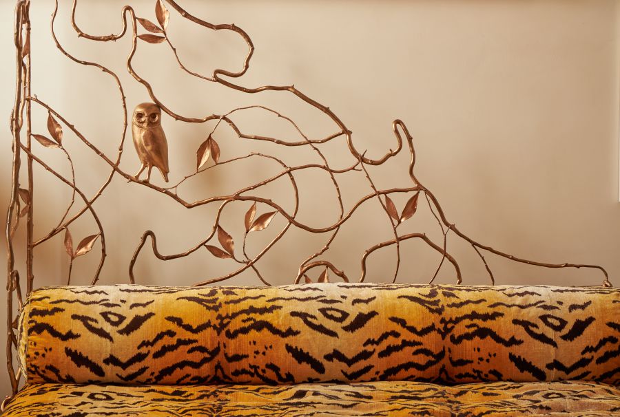Un divano con un motivo a stampa tigrata gialla e nera si trova di fronte a una testata metallica decorativa con rami intrecciati, foglie e un piccolo gufo, che evoca lo stile eclettico di Pauline Karpidas.
