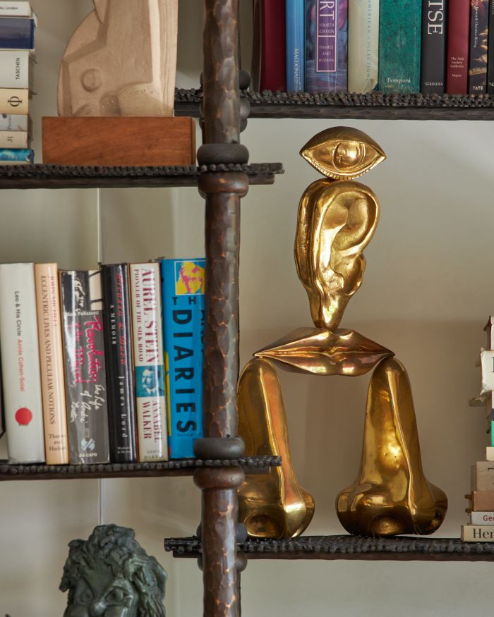 Una scultura astratta in oro lucido con un grande occhio singolo, che ricorda il gusto eclettico di Pauline Karpidas, si trova su uno scaffale tra libri e altri oggetti decorativi in una biblioteca o in uno studio.