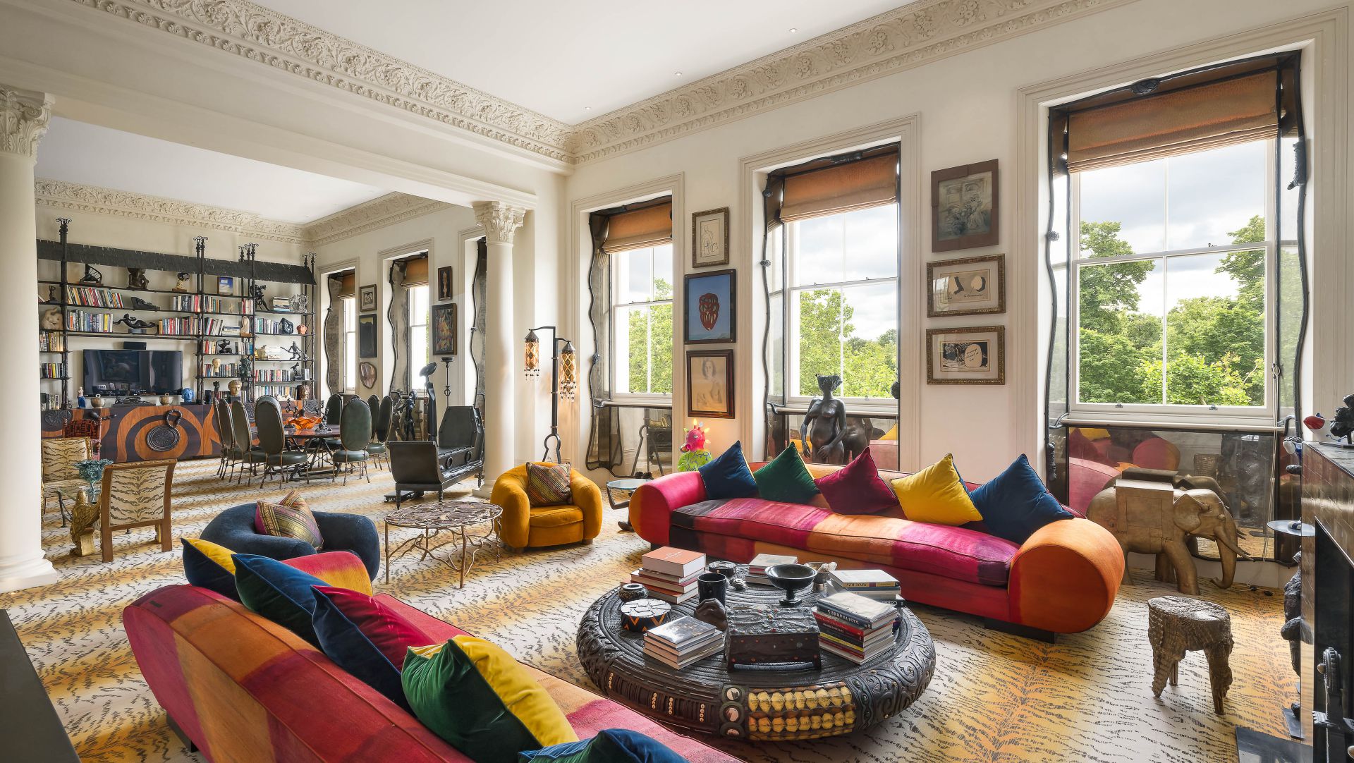 Un soggiorno colorato ed eclettico, ispirato a Pauline Karpidas, è caratterizzato da divani a strisce luminose, cuscini vivaci, grandi finestre con vista sul verde, moquette a motivi, pareti piene di arte, sculture uniche e scaffali di libri sullo sfondo.