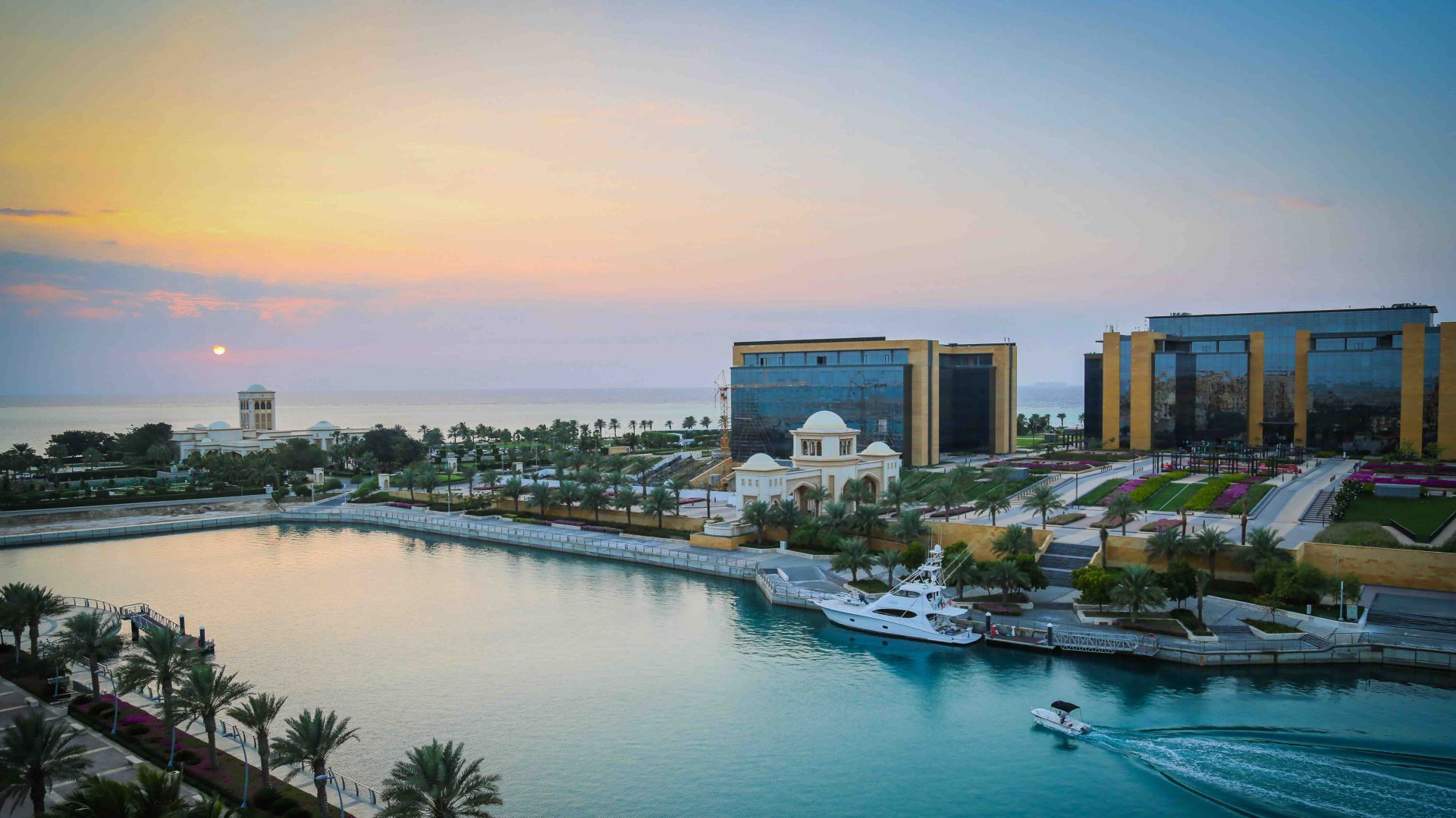 Una veduta del lungomare al tramonto in Arabia Saudita mostra edifici moderni, giardini ordinati, palme, barche e un super yacht su acque calme e turchesi, con il sole che tramonta all'orizzonte e un cielo parzialmente nuvoloso.