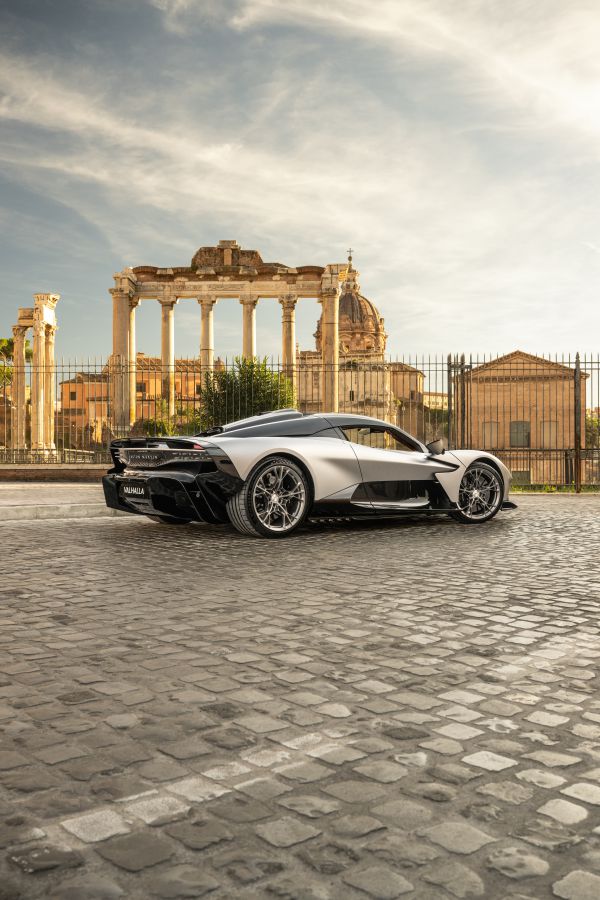 Un'elegante auto sportiva argentata parcheggiata sul selciato di fronte ad antiche rovine romane ed edifici storici sotto un cielo parzialmente nuvoloso.