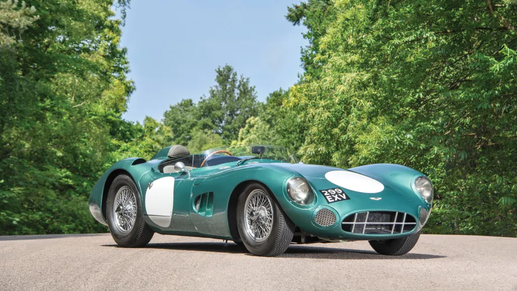 Una classica Aston Martin verde da corsa con tondi bianchi è parcheggiata su una strada circondata da alberi verdeggianti sotto un cielo limpido.