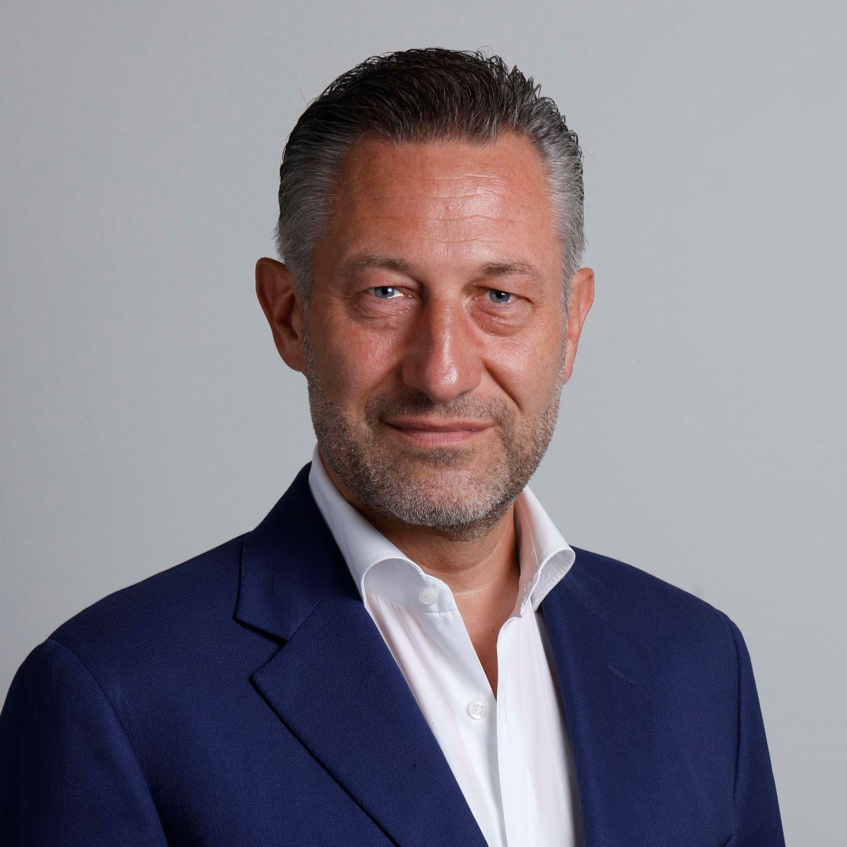 Un uomo di mezza età con capelli corti e lisci e barba chiara indossa una camicia bianca, un blazer blu scuro e un elegante orologio, in piedi su uno sfondo grigio chiaro come se fosse pronto per un concorso di eleganza.