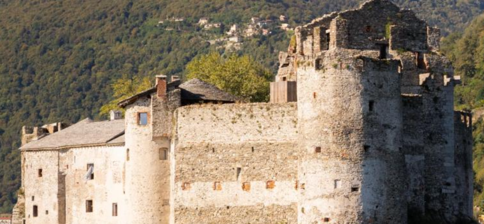 Un antico castello in pietra con torri cilindriche e una parte superiore parzialmente in rovina, che ricorda il Borromeo Kaleon, si erge sullo sfondo di colline boscose e case lontane.