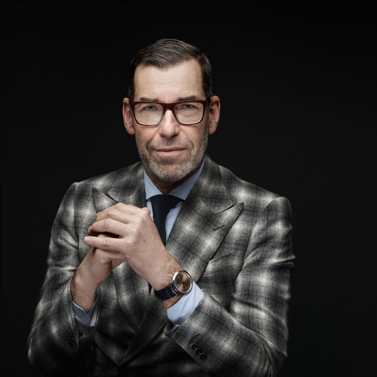 Un uomo con grandi occhiali, abito grigio a quadri, camicia blu e cravatta siede su uno sfondo scuro con le mani giunte e un leggero sorriso: il suo sguardo ricorda lo stile raffinato del concorso eleganza orologi.