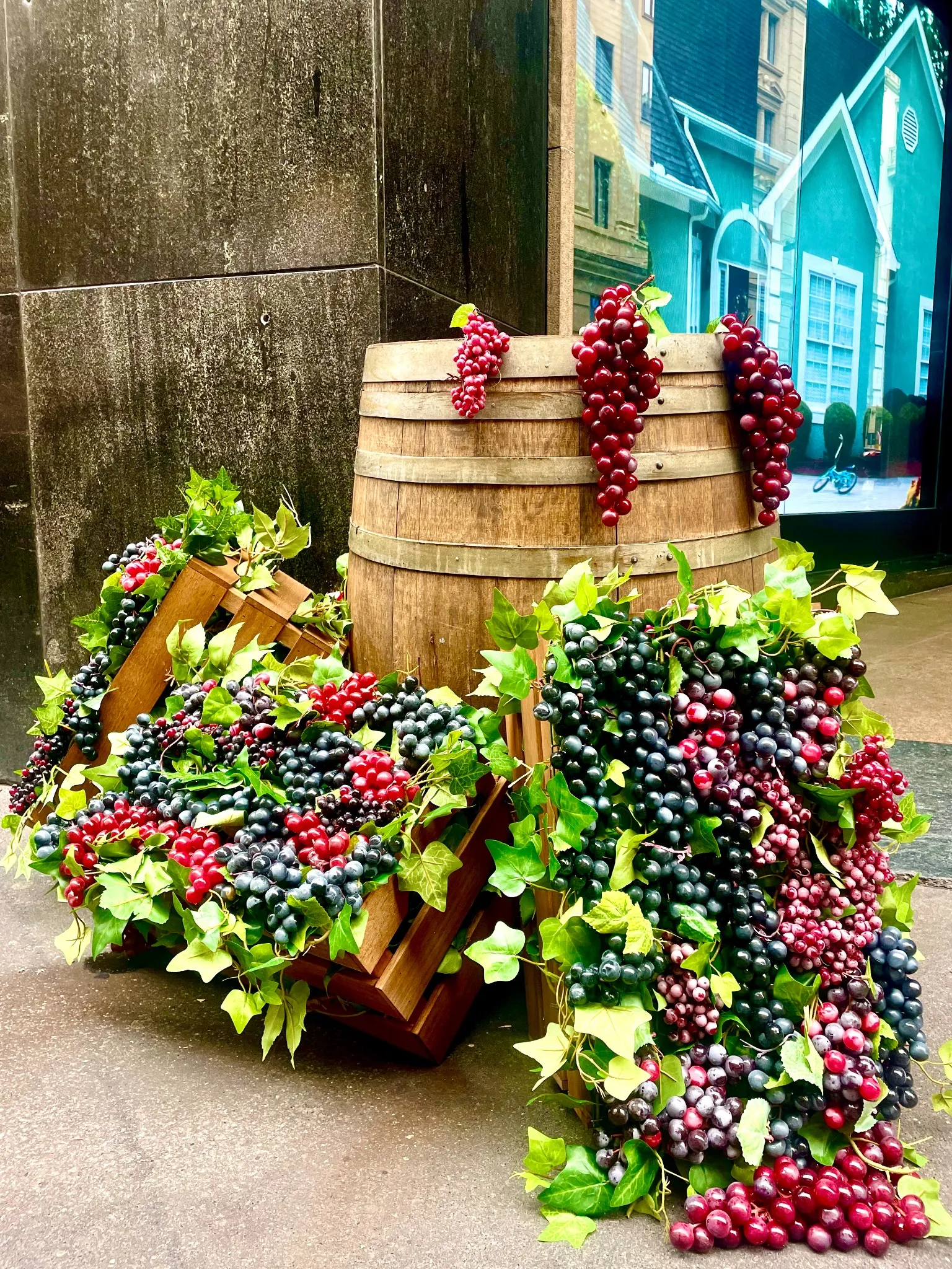 Casse di legno e una botte decorata con grappoli di uva artificiale rossa e viola evocano Vendemmia MonteNapoleone, disposta su un marciapiede cittadino con un edificio e un murale visibili sullo sfondo.
