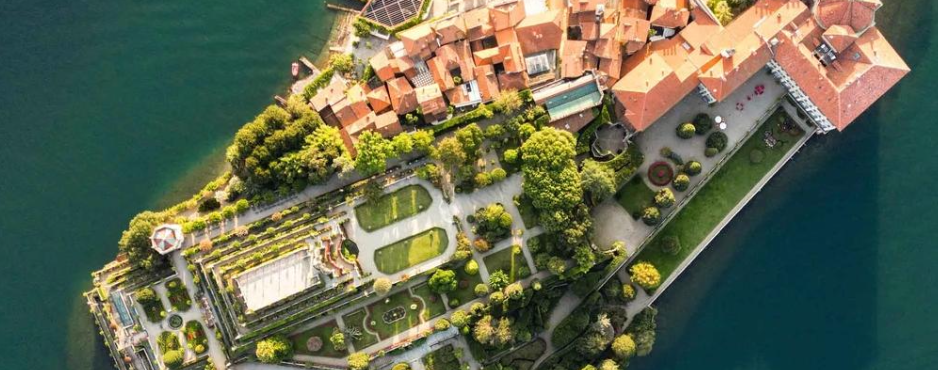 Veduta aerea di una piccola isola con edifici dai tetti rossi e giardini lussureggianti, che ricordano l'eleganza dei Borromeo, circondati da acque verde-blu. Sentieri curati e prati geometrici sono visibili accanto alle strutture.