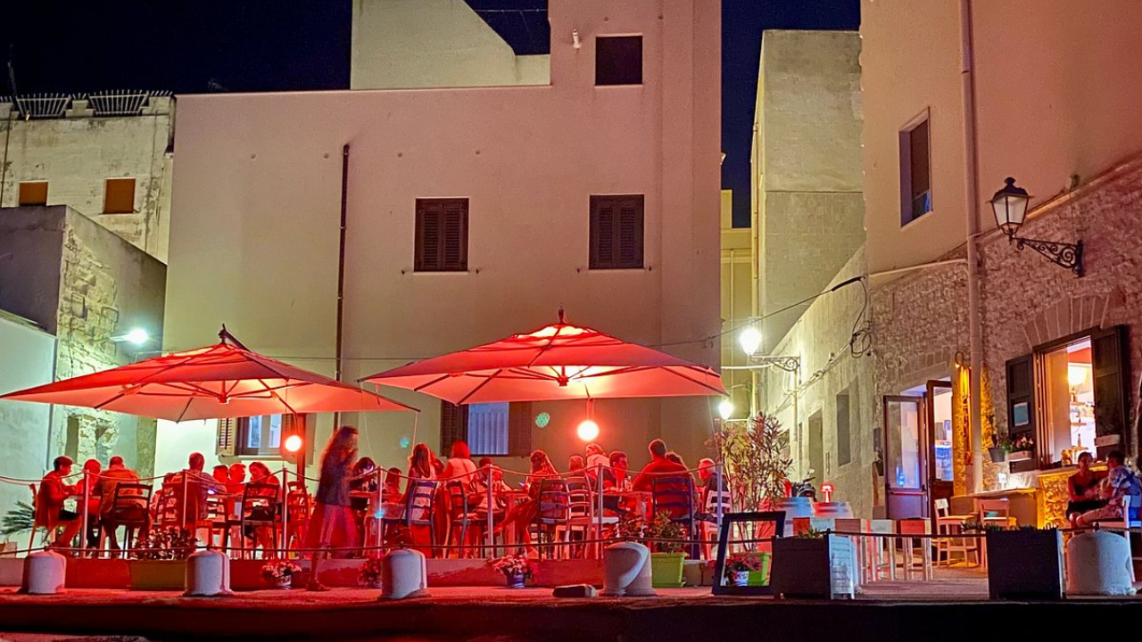 Un ristorante all'aperto di notte a Trapani, con persone che cenano sotto grandi ombrelloni rossi, luci calde ed edifici che circondano il patio. La scena vivace è invitante e piena di fascino siciliano.