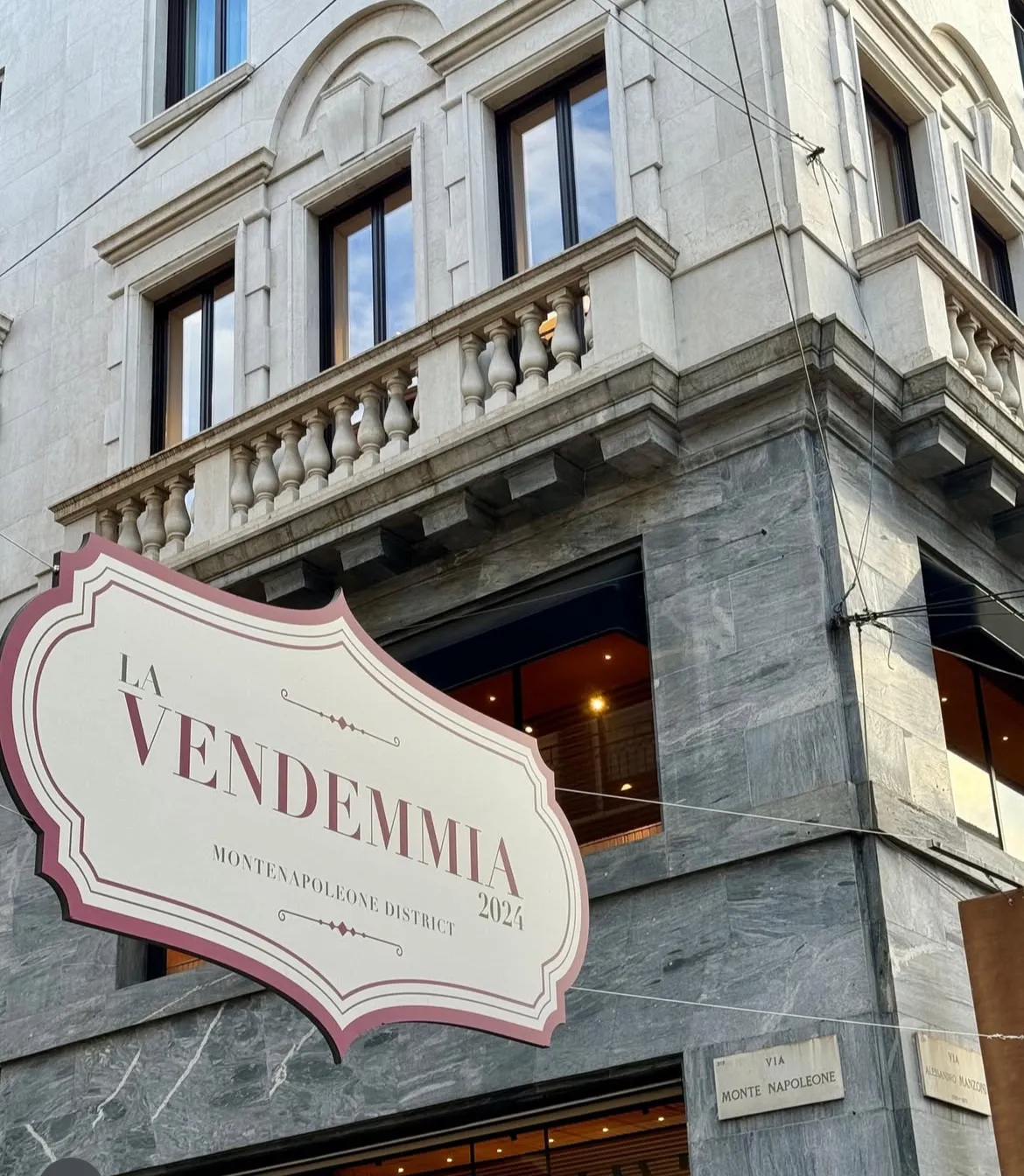 Un grande cartello con la scritta "La Vendemmia 2024 MonteNapoleone District" è appeso a un edificio in pietra con finestre decorate. Un cartello stradale all'angolo recita "Via Monte Napoleone", per celebrare la Vendemmia MonteNapoleone.