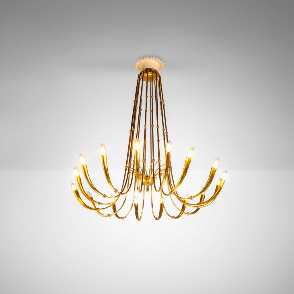 Un moderno lampadario d'oro con dodici bracci ricurvi, ognuno dei quali sostiene una lampadina a forma di candela, pende da un soffitto bianco in una stanza minimale e ben illuminata: un elegante highlight di asta settembre design.