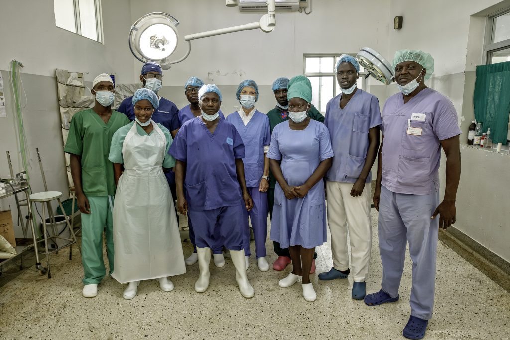 Un gruppo di professionisti del settore medico con camici, mascherine e berretti sta insieme in una sala operatoria di un ospedale, in posa per una foto di gruppo. La stanza è dotata di attrezzature mediche e di un'illuminazione intensa.