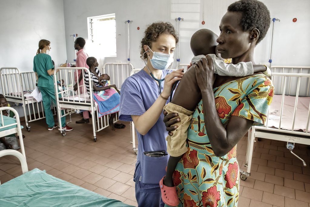 Un operatore sanitario in camice e mascherina esamina un bambino tenuto in braccio da una donna in abiti colorati all'interno di un reparto ospedaliero, con altri pazienti e personale visibili sullo sfondo.