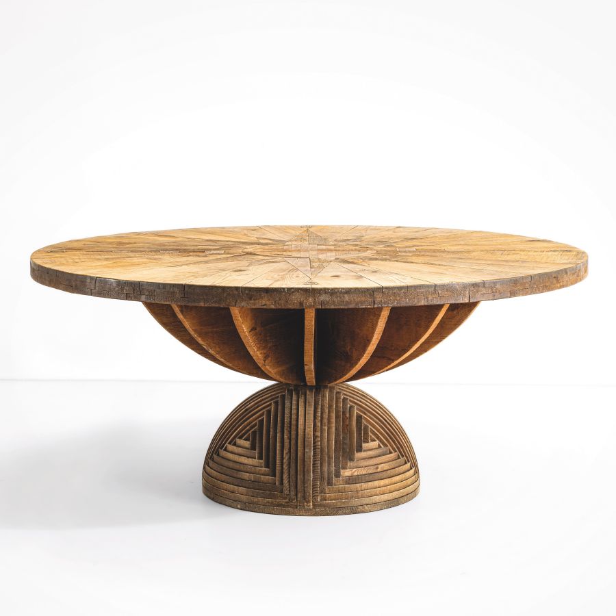 Tavolo rotondo in legno con piano a motivi geometrici e base scultorea, questo pezzo di design unico asta settembre è caratterizzato da pannelli curvi che si intersecano e da una sezione inferiore circolare strutturata, su uno sfondo bianco semplice.