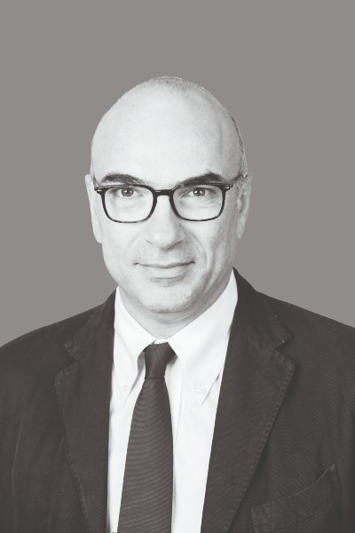 Ritratto in bianco e nero di un uomo con occhiali, giacca, camicia bianca e cravatta su uno sfondo grigio. L'uomo guarda direttamente la macchina fotografica con un'espressione neutra, evocando lo stile classico della fotografia trapanese.